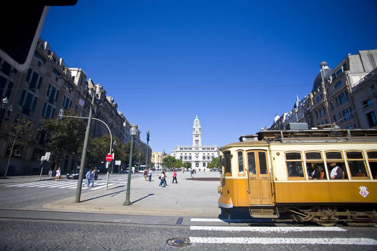 A yellow tram crosses a wide Porto street with the clock tower of City Hall at the far end