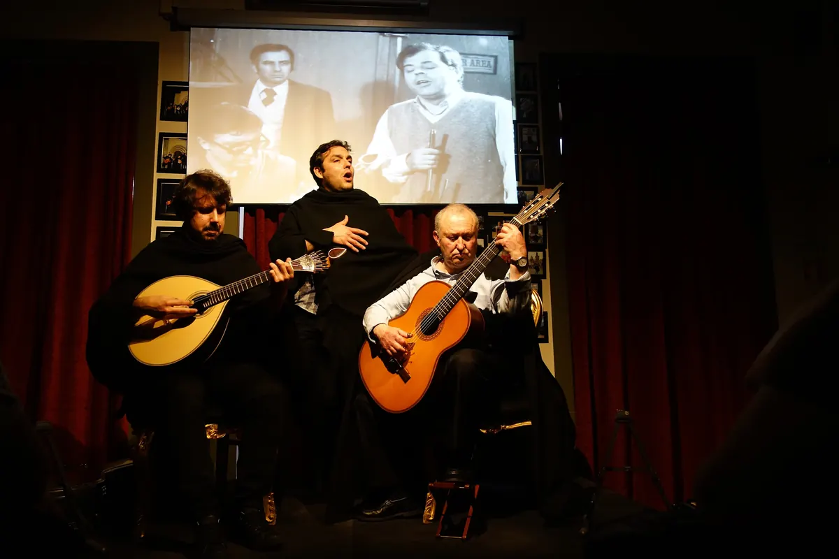 Three men in black capes on a small stage, two with guitars and one singing, in front of red curtains