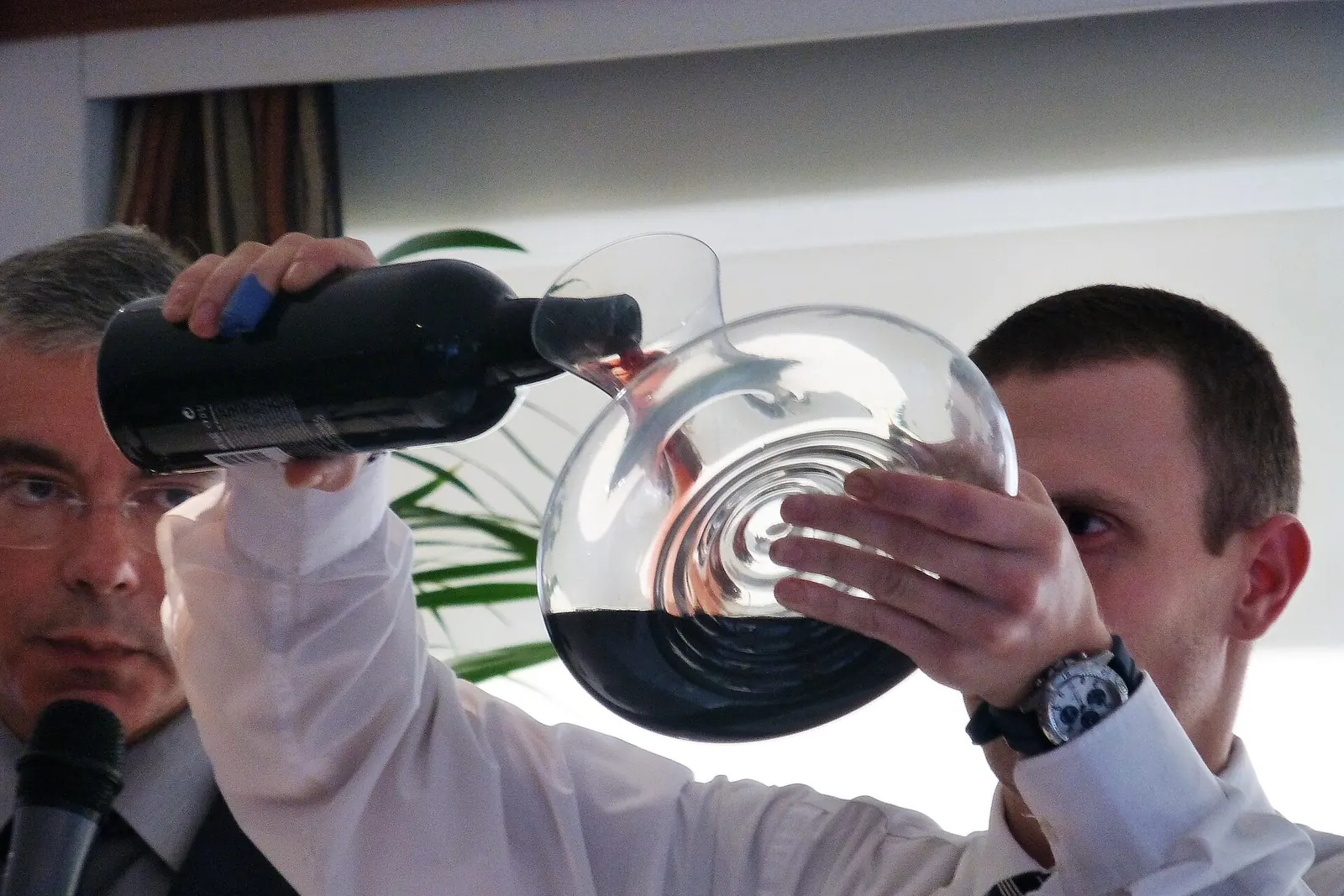 A man tips a dark bottle of port into a glass decanter held up to the light