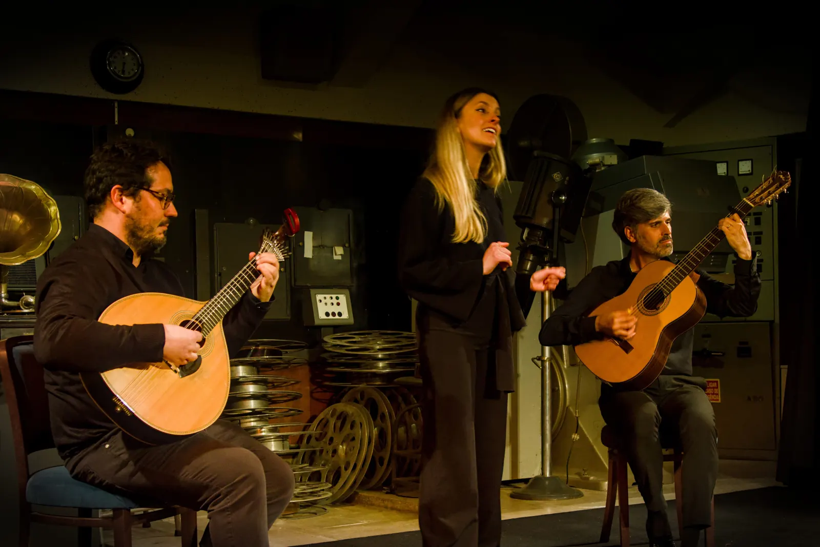 A fado singer between two seated guitarists, one holding a Portuguese guitar and one a classical guitar, on a small stage